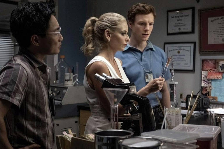 A imagem mostra três pessoas em um ambiente de laboratório. À esquerda, um homem de óculos e camisa listrada observa atentamente. No centro, uma mulher com cabelo preso e vestido branco segura um objeto, enquanto à direita, um homem com cabelo ruivo e camisa azul analisa o que a mulher está segurando. Ao fundo, há prateleiras com frascos e documentos pendurados na parede.