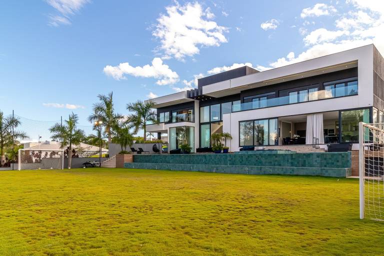 A imagem mostra uma casa moderna de dois andares com grandes janelas e uma fachada em tons de cinza e branco. Na frente, há um amplo gramado verde e bem cuidado, com algumas palmeiras ao redor. À esquerda, há um gol de futebol. O céu está claro com algumas nuvens brancas.