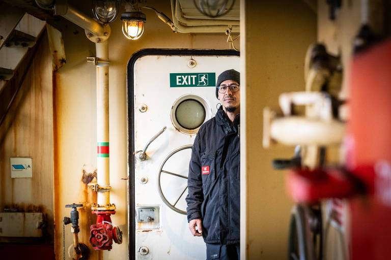 A imagem mostra um homem em um ambiente interno de um navio， posicionado próximo a uma porta de saída. Ele usa um casaco escuro e um gorro， e está olhando para a câmera. Ao fundo， há uma porta com um sinal verde indicando 039;EXIT039; e um visor redondo. O ambiente é composto por paredes metálicas e tubulações visíveis， com uma iluminação suave.