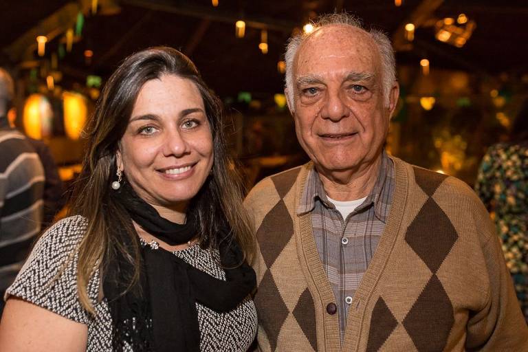 A imagem mostra duas pessoas sorrindo em um ambiente interno. À esquerda, uma mulher com cabelo longo e liso, usando uma blusa cinza e um lenço. À direita, um homem idoso e calvo com cabelo grisalho, vestindo um suéter bege e marrom com padrão de losangos e uma camisa xadrez. O fundo é decorado com luzes e plantas.