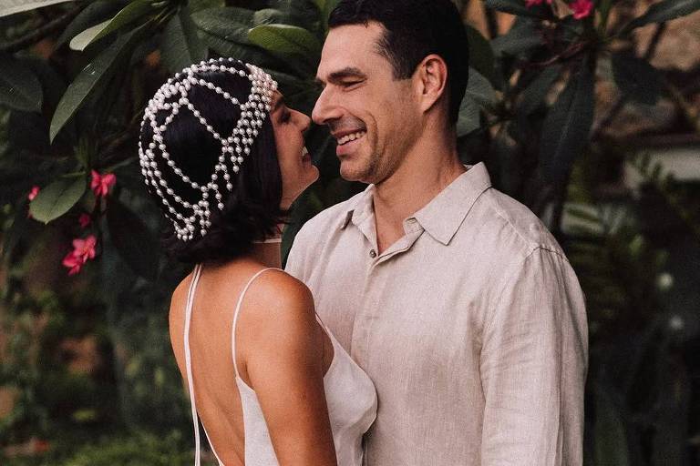 A imagem mostra um casal sorrindo um para o outro em um ambiente natural. A mulher usa um vestido branco com alças finas e uma tiara de pérolas, enquanto o homem veste uma camisa de linho clara. Eles estão em um jardim com plantas verdes e flores rosa ao fundo.