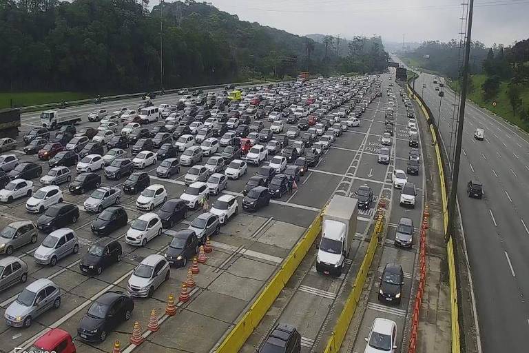 A imagem mostra um grande congestionamento em uma praça de pedágio， com muitos carros parados em várias faixas. À direita， há uma estrada livre， enquanto à esquerda， os veículos estão alinhados， aguardando para passar pelo pedágio. Ao fundo， é possível ver uma área verde e uma estrada que se estende