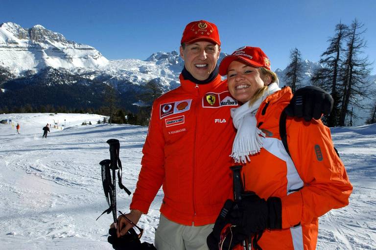 Um casal sorridente posa para a foto em uma estação de esqui. Ambos estão vestidos com roupas de inverno， o homem usa um casaco vermelho， e a mulher， um casaco laranja com um cachecol branco. Ao fundo， há montanhas cobertas de neve e árvores. O céu está claro e ensolarado.