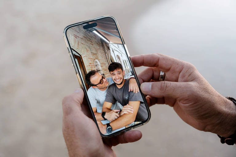 A imagem mostra uma pessoa segurando um celular que exibe uma foto de duas pessoas sorrindo em um ambiente interno. Uma das pessoas está com o braço ao redor da outra， que parece estar fazendo uma expressão engraçada. O fundo da foto no celular apresenta paredes de tijolos expostos e uma decoração simples.