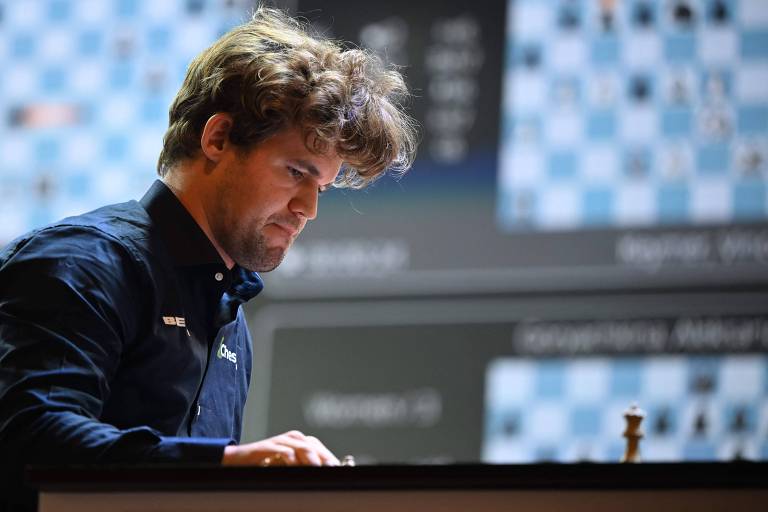 Um jogador de xadrez está concentrado em uma partida， com o olhar voltado para o tabuleiro. Ele tem cabelo cacheado e usa uma camisa escura com o logotipo 039;Chess039;. No fundo， há uma tela mostrando posições de peças de xadrez.
