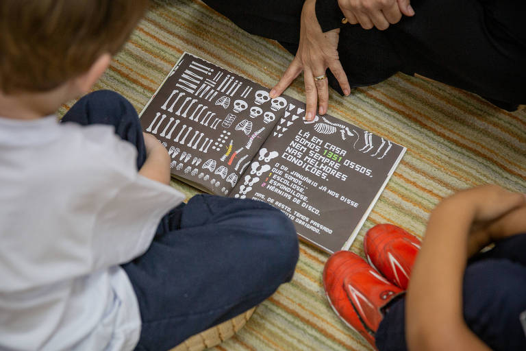 A imagem mostra duas crianças sentadas no chão， observando um livro aberto que está sendo segurado por um adulto. O livro possui um fundo escuro com várias ilustrações e texto em cores claras. As crianças estão vestidas com camisetas claras e calças jeans， uma delas usa tênis vermelhos. O ambiente parece ser uma sala de aula ou um espaço de aprendizado.