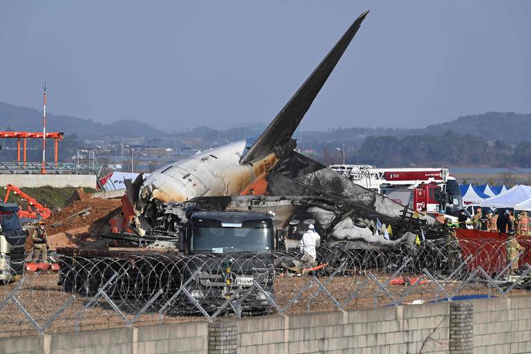 A imagem mostra os destroços de um avião acidentado em um local de pouso. A fuselagem do avião está danificada e parcialmente queimada， com a asa visivelmente quebrada. Há veículos de emergência e equipes de resgate ao redor， além de uma cerca de arame farpado ao fundo. O céu está claro e há montanhas ao longe.