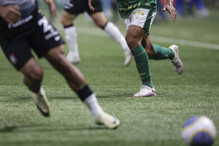 A imagem mostra jogadores de futebol em um campo， com foco nas pernas de um jogador vestindo um uniforme verde e branco， enquanto outro jogador， parcialmente visível， usa um uniforme preto. O campo é coberto de grama artificial e uma bola de futebol está visível ao fundo.
