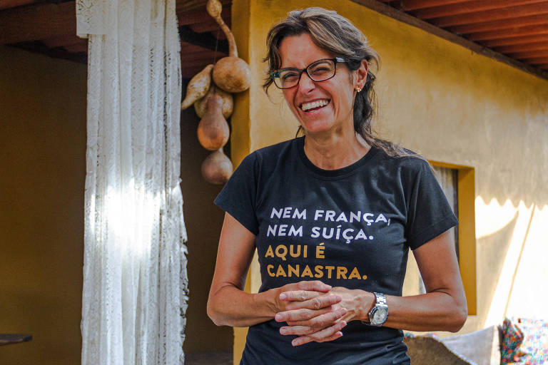 Uma mulher sorridente está em pé, usando uma camiseta preta com a frase 'Nem França, nem Suíça. Aqui é Canastra.' Ela tem cabelos longos e usa óculos. Ao fundo, há uma parede amarela e alguns objetos decorativos pendurados.