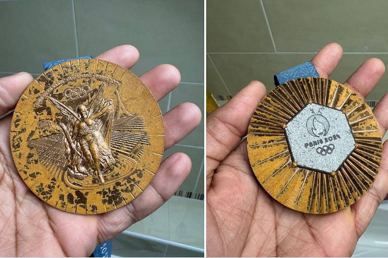 A imagem mostra uma medalha olímpica sendo segurada por uma mão. O lado frontal da medalha apresenta uma figura esculpida de um atleta， com detalhes que incluem os anéis olímpicos e um fundo com linhas radiantes. O verso da medalha é hexagonal， com um design em relevo que inclui a inscrição 039;PARIS 2024039; e os anéis olímpicos. A medalha é de cor dourada e possui uma fita azul.