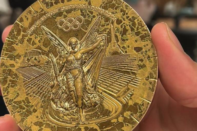 A imagem mostra uma medalha olímpica dourada sendo segurada por uma mão. A medalha apresenta uma figura central de uma mulher alada， simbolizando a vitória， com detalhes de um estádio ao fundo. Na parte superior， estão os anéis olímpicos. Ao fundo， há uma pessoa desfocada e um ambiente interno com iluminação suave.