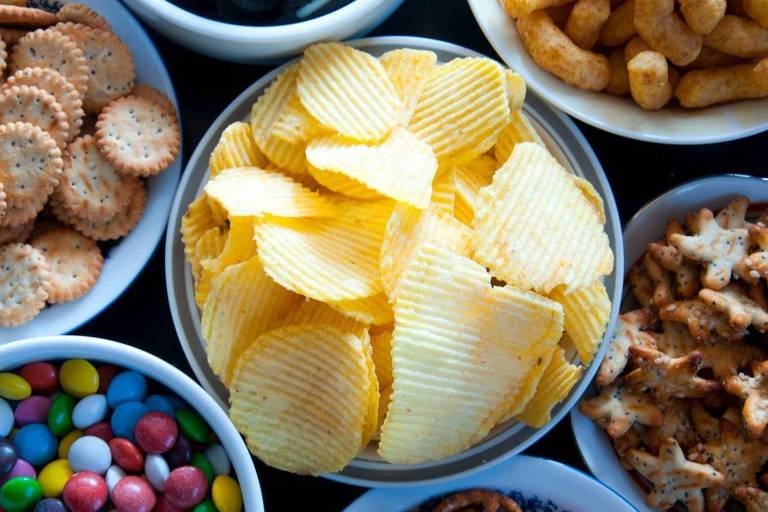 A imagem apresenta uma variedade de petiscos dispostos em tigelas. No centro， uma tigela com batatas chips amarelas onduladas domina a composição. Ao redor， há tigelas com diferentes lanches， incluindo bolachas salgadas redondas， balas de chocolate coloridas， salgadinhos dourados e um mix de castanhas e frutas secas. A cena é vibrante， com um enfoque no contraste das cores e texturas dos alimentos.