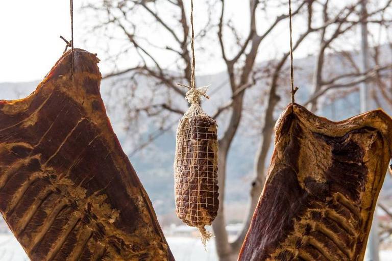 A imagem mostra cortes de carne seca pendurados ao ar livre， aparentemente em um ambiente rural. Há pedaços grandes de carne， com as costelas visíveis， suspensos em estruturas ou cordas. No centro， também está pendurado um embutido， como salame ou linguiça curada， envolto em uma malha fina. Ao fundo， há árvores sem folhas， indicando uma estação fria， e montanhas ao longe， compondo um cenário natural. A luz é suave， sugerindo um dia nublado ou de inverno.