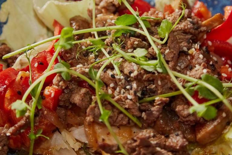 Carne frita com gergelim e verduras em um prato azul em close-up， beshbarmak em um prato com gergelim e verduras， carne cozida no estilo oriental.
