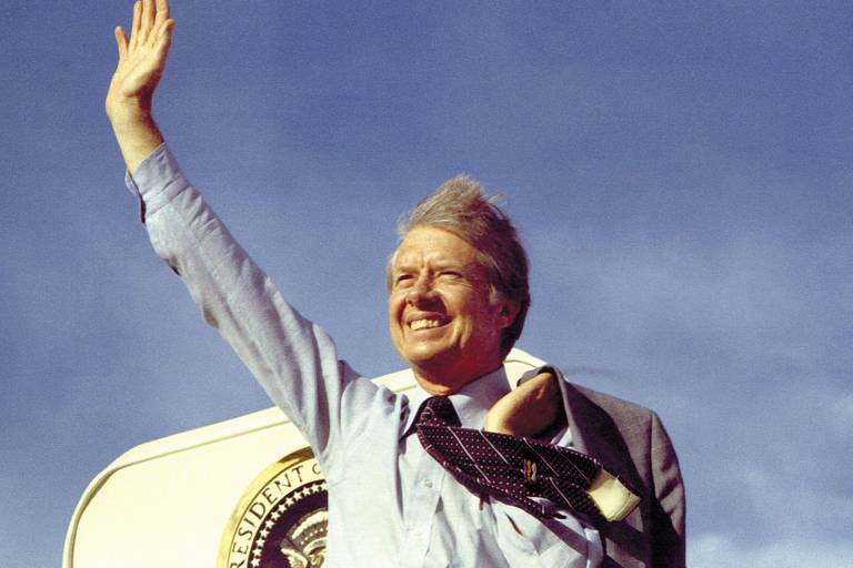 Um homem está acenando com a mão direita, sorrindo, em frente a um fundo de céu azul. Ele usa uma camisa azul e uma gravata escura. Ao fundo, é visível um símbolo presidencial.