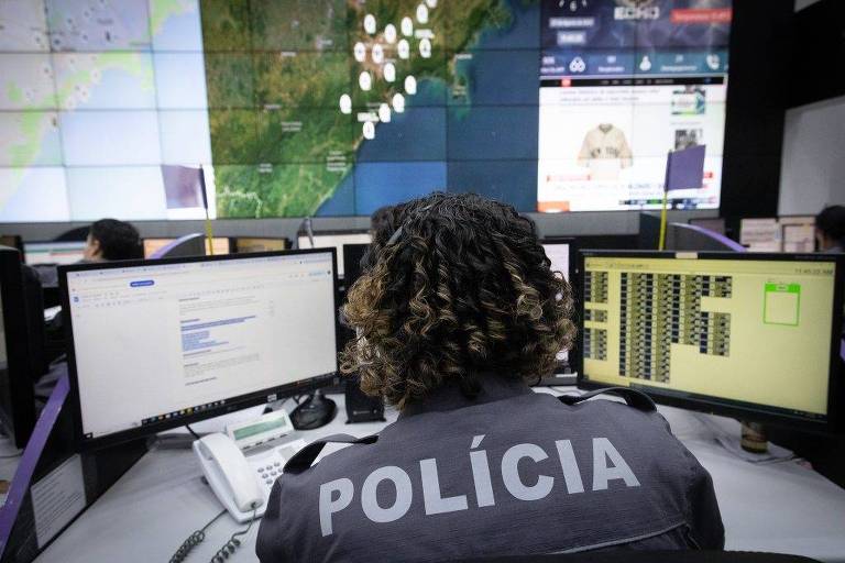 A imagem mostra uma sala de controle policial com várias telas exibindo informações. Uma pessoa， vista de costas， usa uma camisa com a palavra 039;POLÍCIA039; em destaque. Ao fundo， há um grande painel com um mapa e dados visuais， enquanto outras pessoas trabalham em estações de computador.