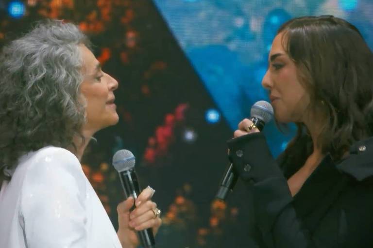 Em foto colorida, duas mulheres aparecem cantando no palco