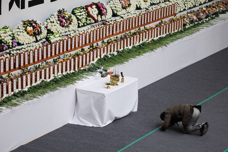 A imagem mostra uma cerimônia de luto com uma longa fileira de flores brancas e arranjos florais coloridos dispostos em uma parede. Na parte inferior da imagem, um homem está ajoelhado em frente a uma mesa coberta com uma toalha branca, que contém alguns objetos, incluindo garrafas. O ambiente parece ser um espaço formal, possivelmente um salão de eventos.