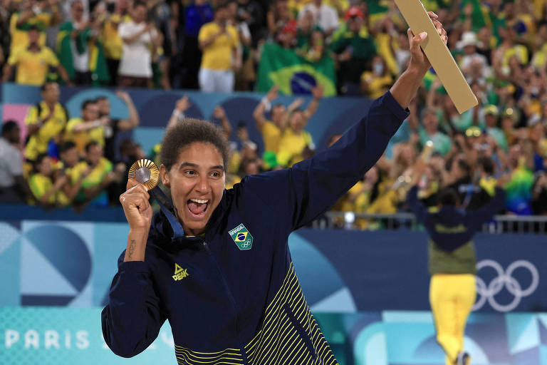 Uma atleta brasileira está sorrindo e segurando uma medalha em uma mão e um bastão na outra, celebrando sua vitória. Ao fundo, uma multidão vestida com roupas nas cores da bandeira do Brasil, em um ambiente festivo, provavelmente durante os Jogos Olímpicos.