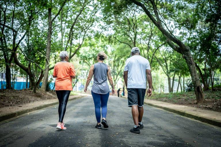 A imagem mostra três pessoas caminhando em uma estrada cercada por árvores. A primeira pessoa， à esquerda， usa uma camiseta laranja e calças pretas. A segunda pessoa， no meio， está vestindo uma blusa cinza e calças azuis. A terceira pessoa， à direita， usa uma camiseta branca e calças escuras. O ambiente é natural， com muita vegetação ao redor.