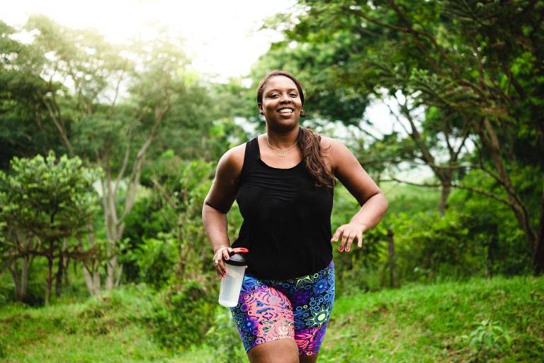 Mulher negra caminhando， sorrindo， com uma garrafa de água na mão. Ao fundo， árvores e gramado.
