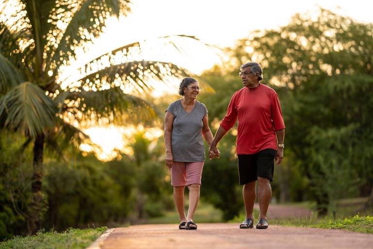 Um casal de idosos caminha de mãos dadas em um caminho cercado por árvores e palmeiras， durante o pôr do sol. A mulher usa uma camiseta cinza e shorts， enquanto o homem veste uma camiseta vermelha e shorts pretos. O ambiente é tranquilo e natural， com uma luz suave do sol ao fundo.