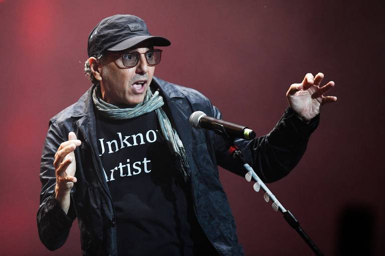 Um homem em um palco, usando óculos escuros e um boné, está se apresentando com um microfone. Ele veste uma camiseta preta com a frase 'Unknown Artist' e uma jaqueta escura. O fundo é desfocado, com iluminação em tons de vermelho.