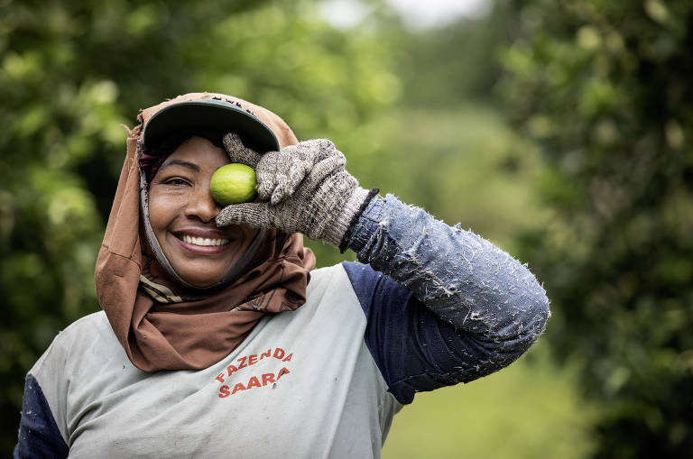 Uma mulher sorridente segura um limão em frente ao rosto, usando luvas e um chapéu. Ela está vestida com uma camiseta que diz 'Fazenda Sara'. Ao fundo, há uma paisagem verde com árvores.