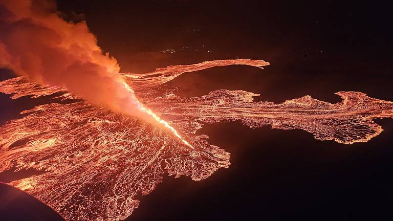 A imagem mostra uma erupção vulcânica noturna, com lava fluindo em um padrão sinuoso. Há uma coluna de fumaça e cinzas sendo expelida do vulcão, iluminando a área ao redor com um brilho laranja intenso. O cenário é dramático, destacando a força da natureza.