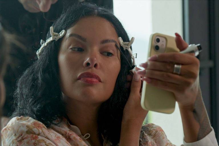 A imagem mostra uma mulher com cabelo cacheado e escuro, usando prendedores brancos no cabelo. Ela está olhando para um celular que segura com a mão direita, enquanto com a mão esquerda ajusta o cabelo. A mulher tem lábios vermelhos e está vestindo uma blusa com estampa floral. Ao fundo, há uma pessoa que parece estar ajudando com a maquiagem.