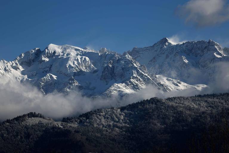 A imagem mostra uma cadeia de montanhas cobertas de neve sob um céu azul. As montanhas têm picos agudos e algumas nuvens brancas estão visíveis na parte inferior， enquanto a vegetação escura é visível na base das montanhas.