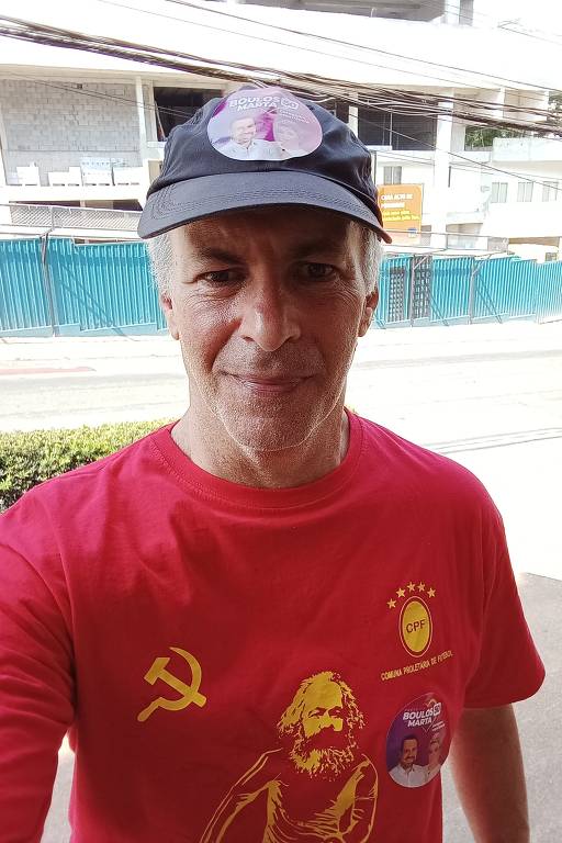 Um homem está posando para a foto, usando uma camiseta vermelha com uma imagem de Karl Marx e um símbolo comunista. Ele também está usando um boné preto com um adesivo que diz 'BOULOS 50'. O fundo mostra uma construção e uma cerca azul.