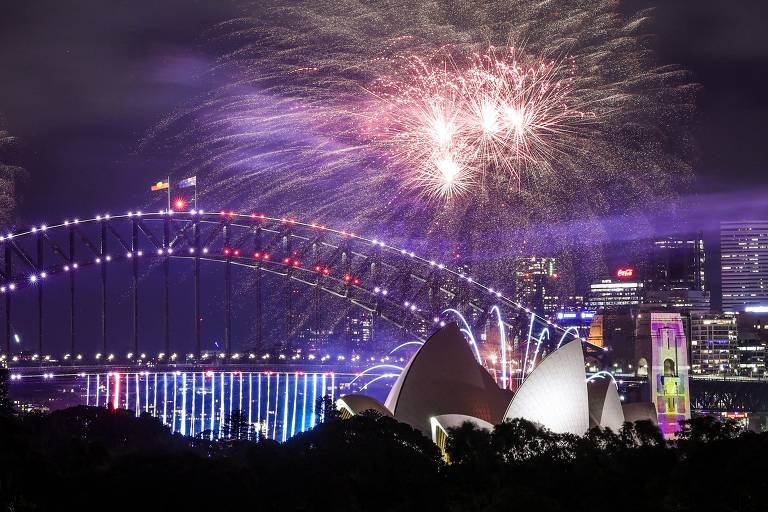 A imagem mostra uma vista noturna da Sydney Harbour Bridge e da Sydney Opera House, iluminadas com luzes coloridas. Fogos de artifício explodem no céu, criando um espetáculo visual. O cenário urbano ao fundo é visível, com edifícios iluminados.