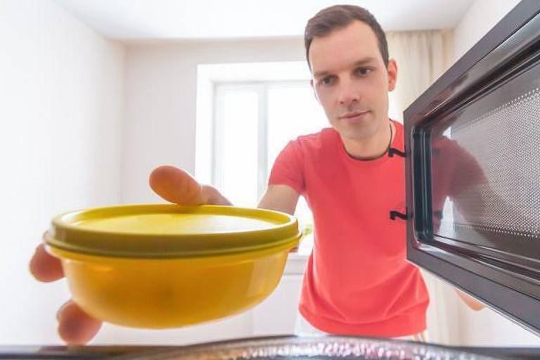 Foto mostra um homem branco， com cabelos castanhos curtos， vestindo uma camiseta vermelha abrindo a porta de um micro-ondas com a mão esquerda e colocando， dentro do aparelho， um pote de plástico amarelo com a mão direita. Temos a visão de dentro do micro-ondas. 