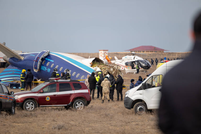 A imagem mostra o local de um acidente aéreo， onde um avião está tombado em um campo. Há várias pessoas， incluindo equipes de resgate e autoridades， trabalhando na cena. Veículos de emergência estão presentes， e um helicóptero pode ser visto ao fundo. O céu está claro e a área ao redor é desértica.