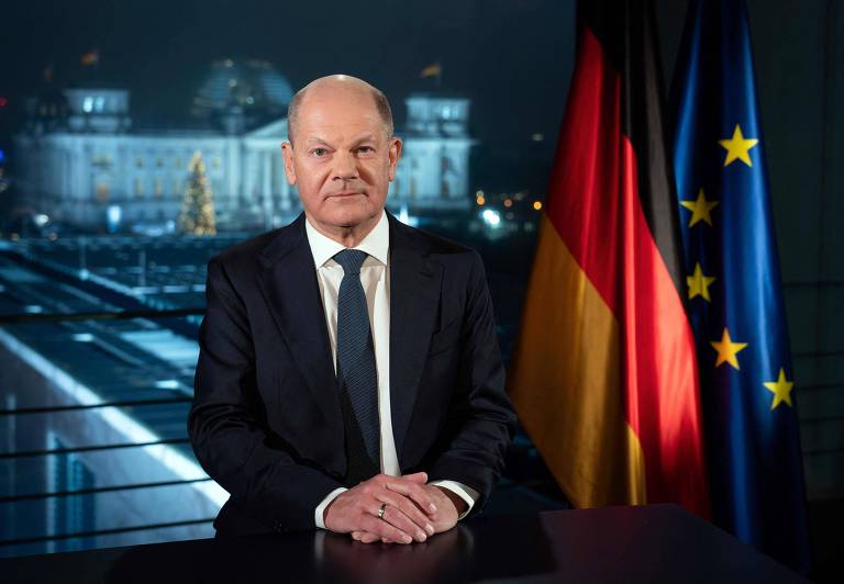 Um homem em um terno escuro está sentado à mesa， com um fundo que mostra o edifício do Reichstag iluminado à noite. Ele tem um sorriso leve e está posicionado entre as bandeiras da Alemanha e da União Europeia.