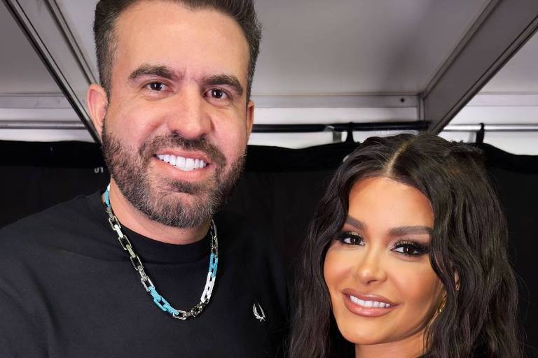 A imagem mostra duas pessoas posando juntas. À esquerda, um homem com cabelo curto e barba, vestindo uma camiseta preta e um colar. À direita, uma mulher com cabelo longo e ondulado, usando um vestido branco com detalhes metálicos. Eles estão sorrindo e a mulher está com a mão sobre a mão do homem.