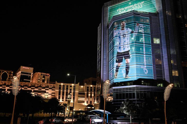 A imagem mostra um grande painel publicitário iluminado à noite， exibindo a imagem de um jogador de futebol em uma camisa branca e preta. O painel está localizado em um edifício alto， com luzes da cidade ao fundo， incluindo edifícios iluminados e uma atmosfera urbana.