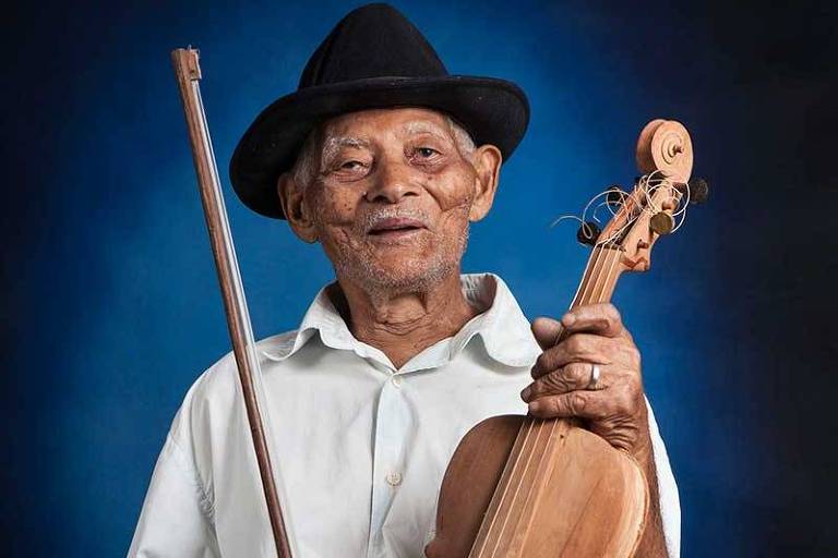 Um homem idoso está sentado, segurando um violino em uma mão e um arco na outra. Ele usa um chapéu preto e uma camisa branca. O fundo é de cor azul escura, destacando sua figura.