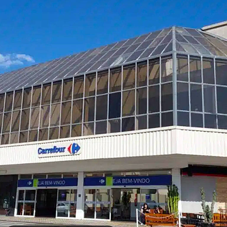 A imagem mostra a fachada de um supermercado Carrefour. A estrutura é moderna, com uma grande área envidraçada no andar superior e uma entrada ampla no térreo. Abaixo da área envidraçada, há uma faixa azul com o texto 'SEJA BEM-VINDO' em letras brancas. O céu está claro e azul ao fundo.