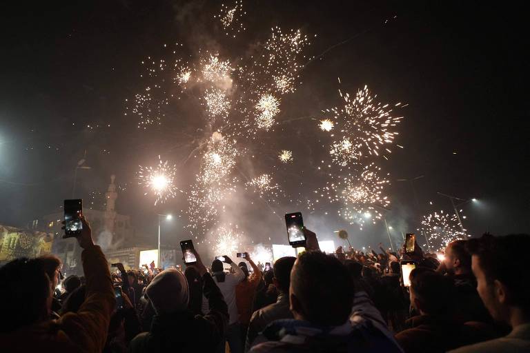 A imagem mostra uma multidão de pessoas em uma celebração noturna， levantando seus celulares para registrar o momento. No fundo， há uma explosão de fogos de artifício iluminando o céu， criando um espetáculo de luzes. A cena é vibrante e festiva， com a presença de edifícios ao fundo， que não são claramente identificáveis.