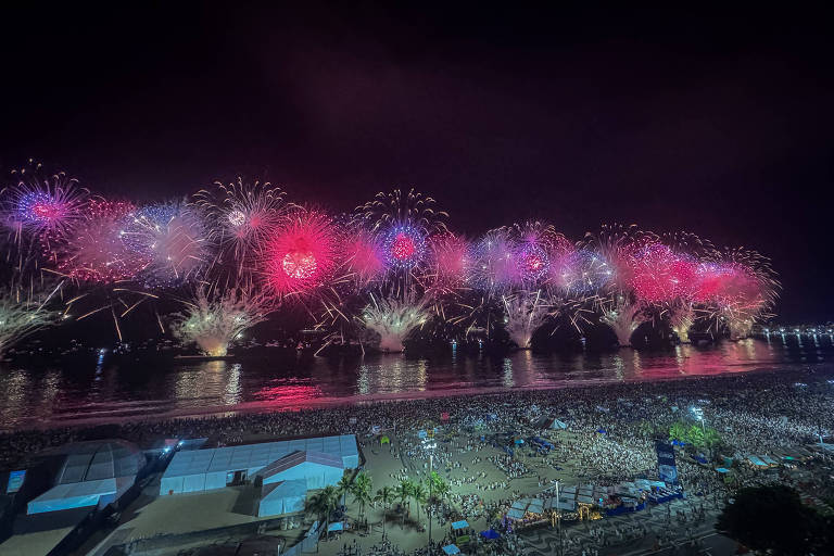 A imagem mostra um espetáculo de fogos de artifício iluminando o céu noturno， com explosões de cores como rosa e roxo. A cena é refletida na água， e uma grande multidão pode ser vista na praia， observando o show. Estruturas e barracas estão visíveis na parte inferior da imagem.