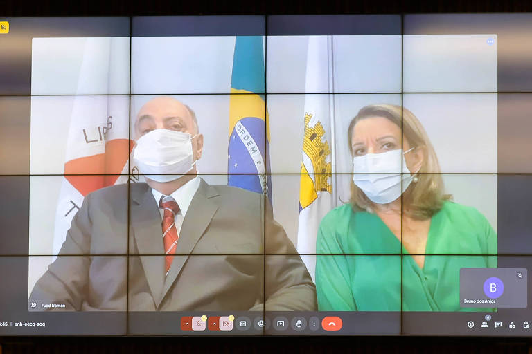 A imagem mostra uma videoconferência em um grande painel. Dois indivíduos estão visíveis: um homem à esquerda， usando um terno escuro e gravata vermelha， e uma mulher à direita， vestindo uma blusa verde. Ambos estão usando máscaras faciais. Ao fundo， há bandeiras do Brasil e de uma cidade， além de um logotipo visível. Na parte inferior da tela， há ícones de controle de videoconferência.