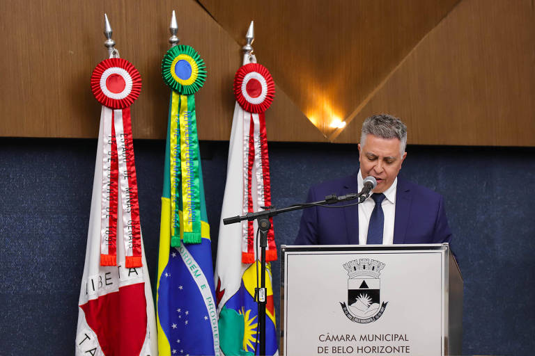 Um homem em um terno escuro está falando em um microfone em frente a um púlpito. Ao fundo, há três bandeiras: uma do Brasil, uma de Minas Gerais e uma de Belo Horizonte. O ambiente é formal, com uma parede azul escura e detalhes em madeira.