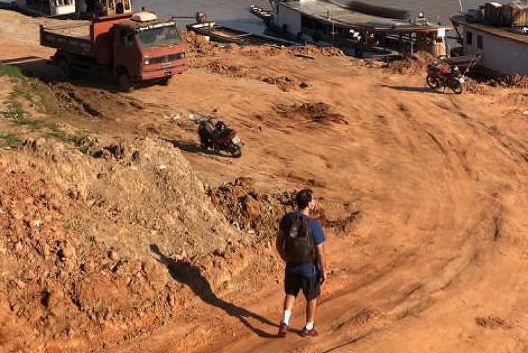 A imagem mostra uma paisagem de um rio com várias embarcações ancoradas na margem. Há uma pessoa caminhando na areia ao primeiro plano， e ao fundo， pode-se ver vegetação e montanhas sob um céu azul com algumas nuvens.