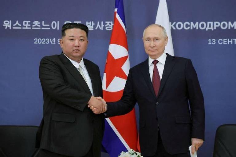 Foto mostra Kim e Putin apertando as mãos na frente da bandeira dos dois países