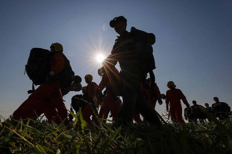 Foto mostra um grupo de soldados， cobertos por penumbras pois a imagem está contra o sol， caminhando em uma gramado