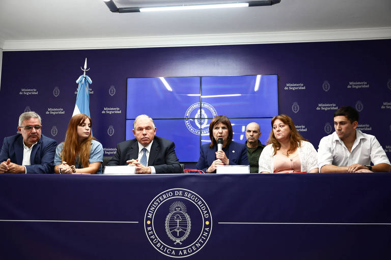 A imagem mostra uma mesa com seis pessoas sentadas， em um ambiente de conferência. Ao fundo， há uma tela com um logotipo e a bandeira da Argentina. A pessoa no centro está falando ao microfone， enquanto os outros escutam atentamente. O ambiente é bem iluminado e decorado com cores sóbrias.
