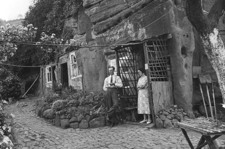 Uma foto em preto e branco de um homem e uma mulher na entrada de uma casa de pedra， com a varanda de madeira encostada em uma caverna de arenito com janelas e portas cortadas. Ele usa camisa e gravata brancas e calças com suspensórios. Ela está usando um vestido claro com uma estampa. Elas estão em um caminho de tijolos do lado de fora da casa. Casas de pedra em Kinver.
