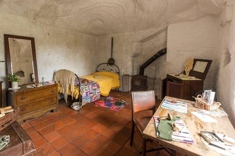 Interior de uma casa com azulejos avermelhados no chão. Uma cama de ferro fica em um canto com uma cômoda de madeira escura ao lado， com um espelho em cima. Outros móveis， incluindo uma mesa e uma cadeira， estão no quarto. Um cano de um fogão a lenha pode ser visto ao lado da cama.
